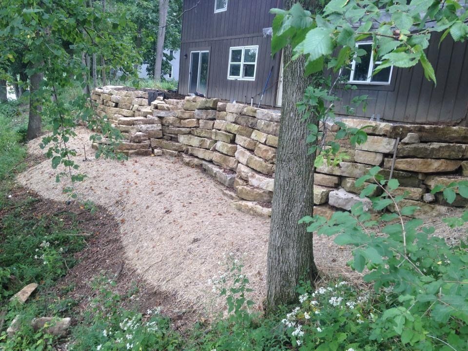 A stone wall is surrounded by trees and grass in front of a house.