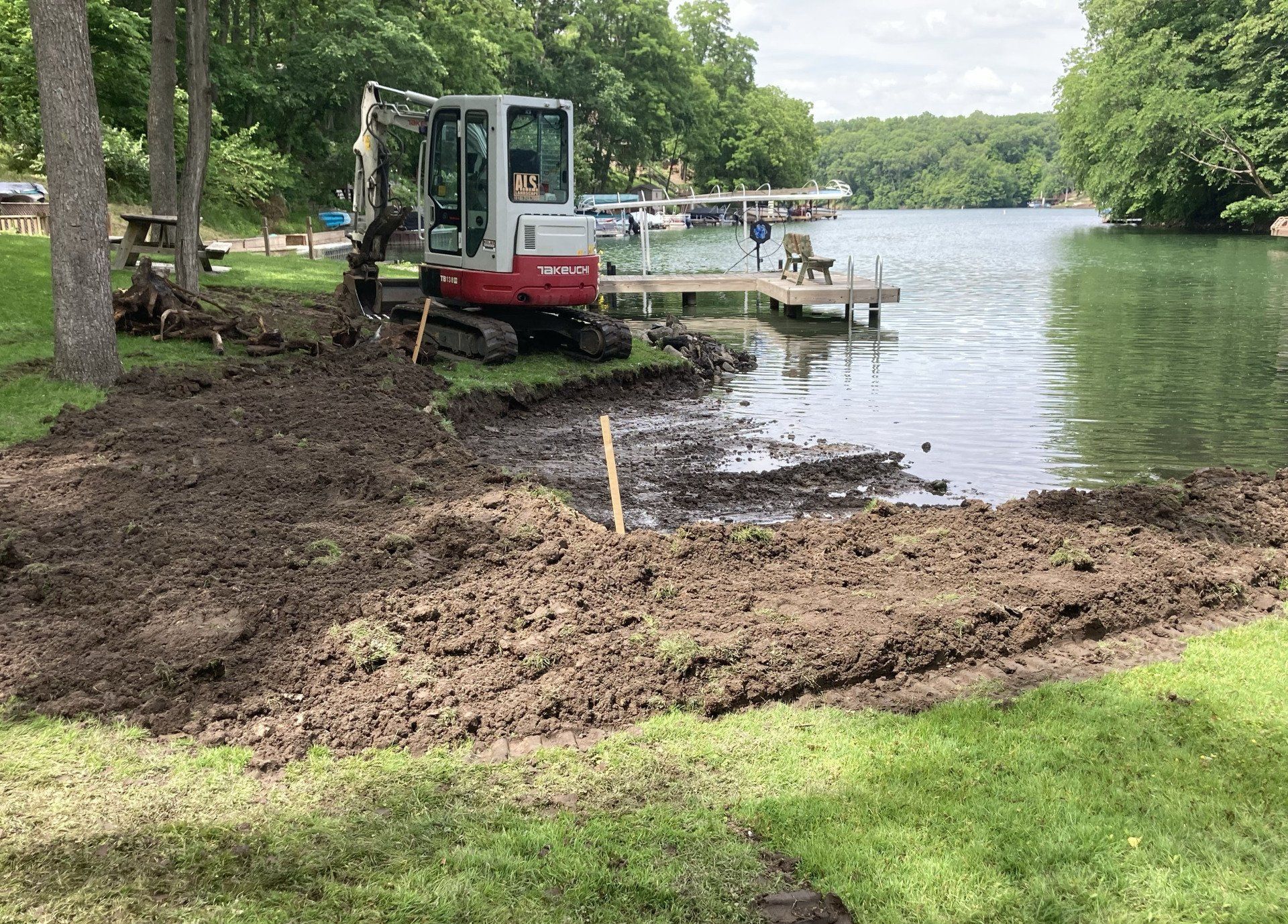 A small excavator is digging a hole in the grass next to a lake.
