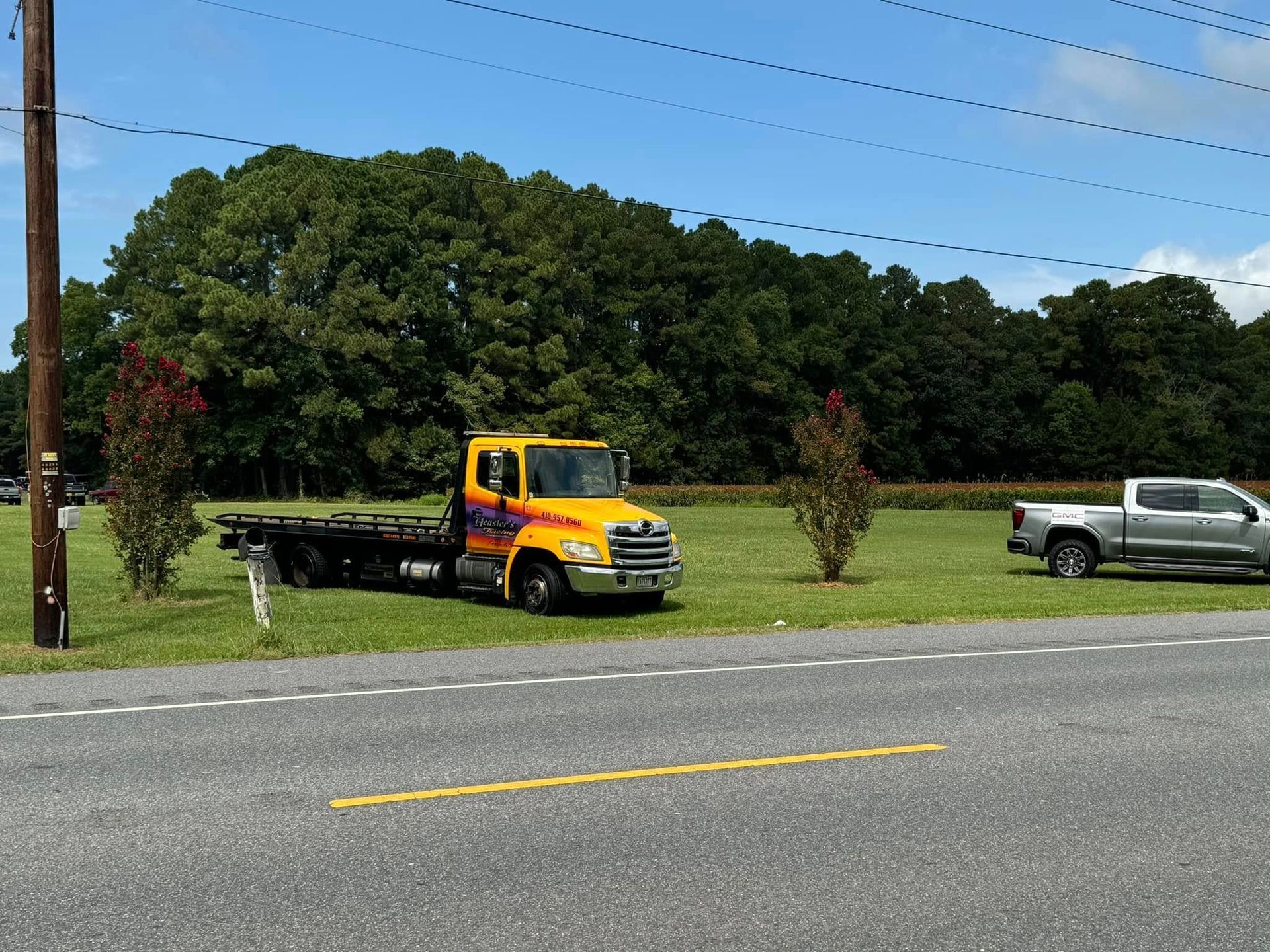 Yellow tow truck parked on grass next to a road, silver pickup truck nearby, trees in the background under a blue sky.