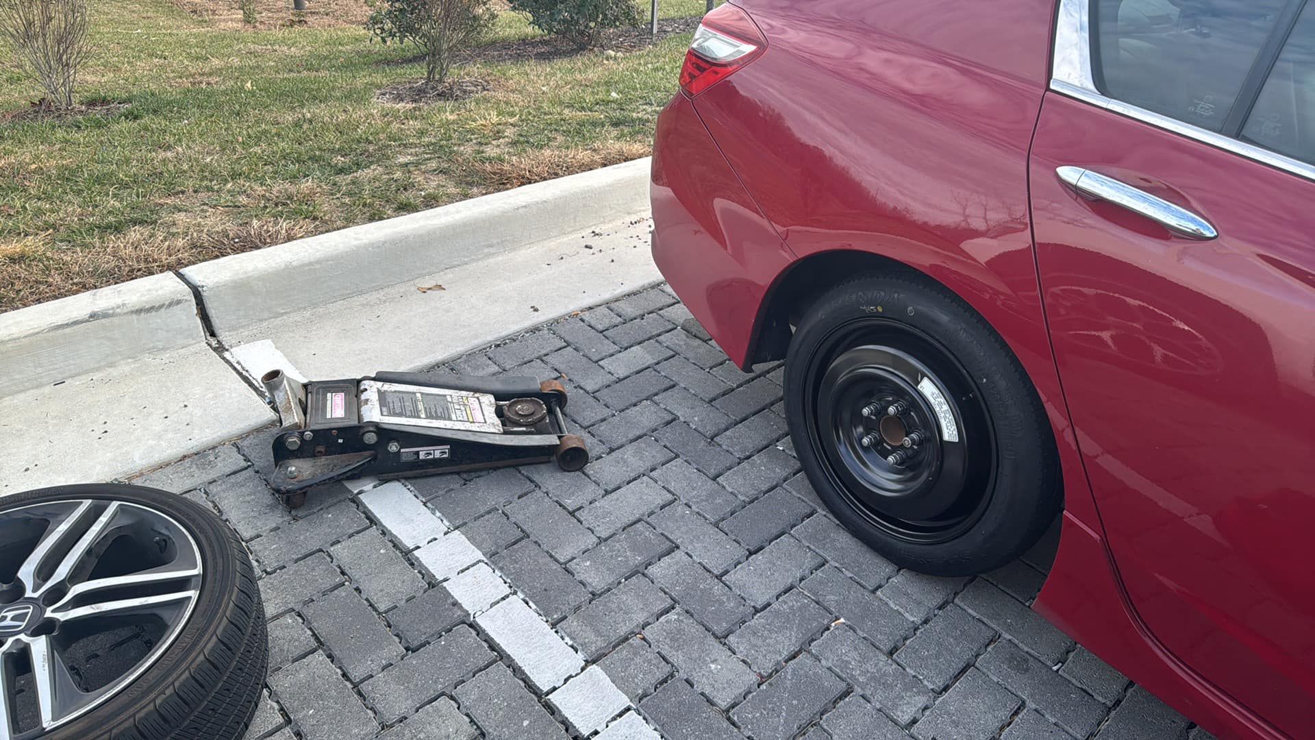 Red car with a flat tire, jack, and spare tire on a brick parking lot.