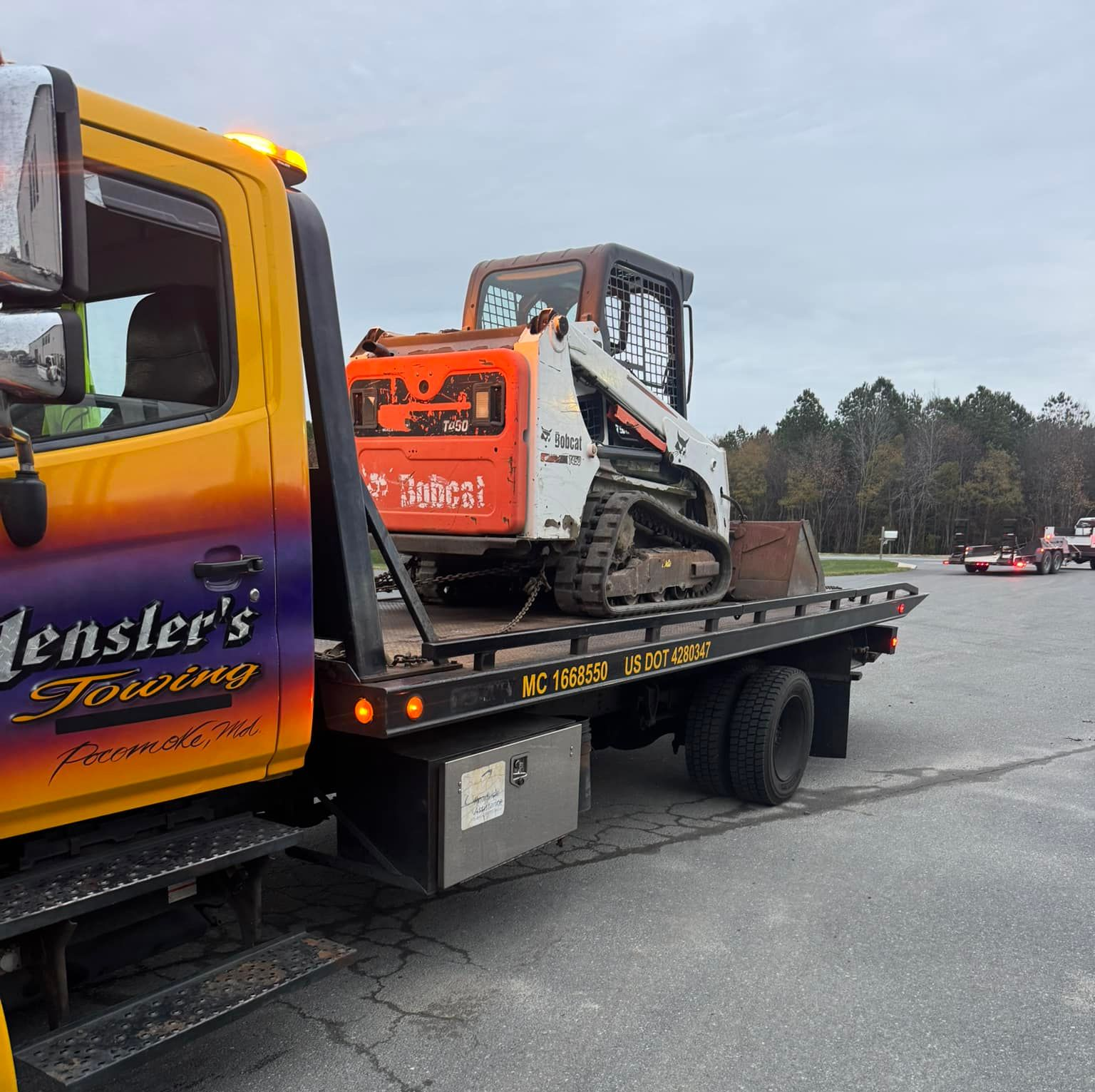 Yellow tow truck carrying a white Jeep Wagoner on a gravel lot; a red car is parked nearby.