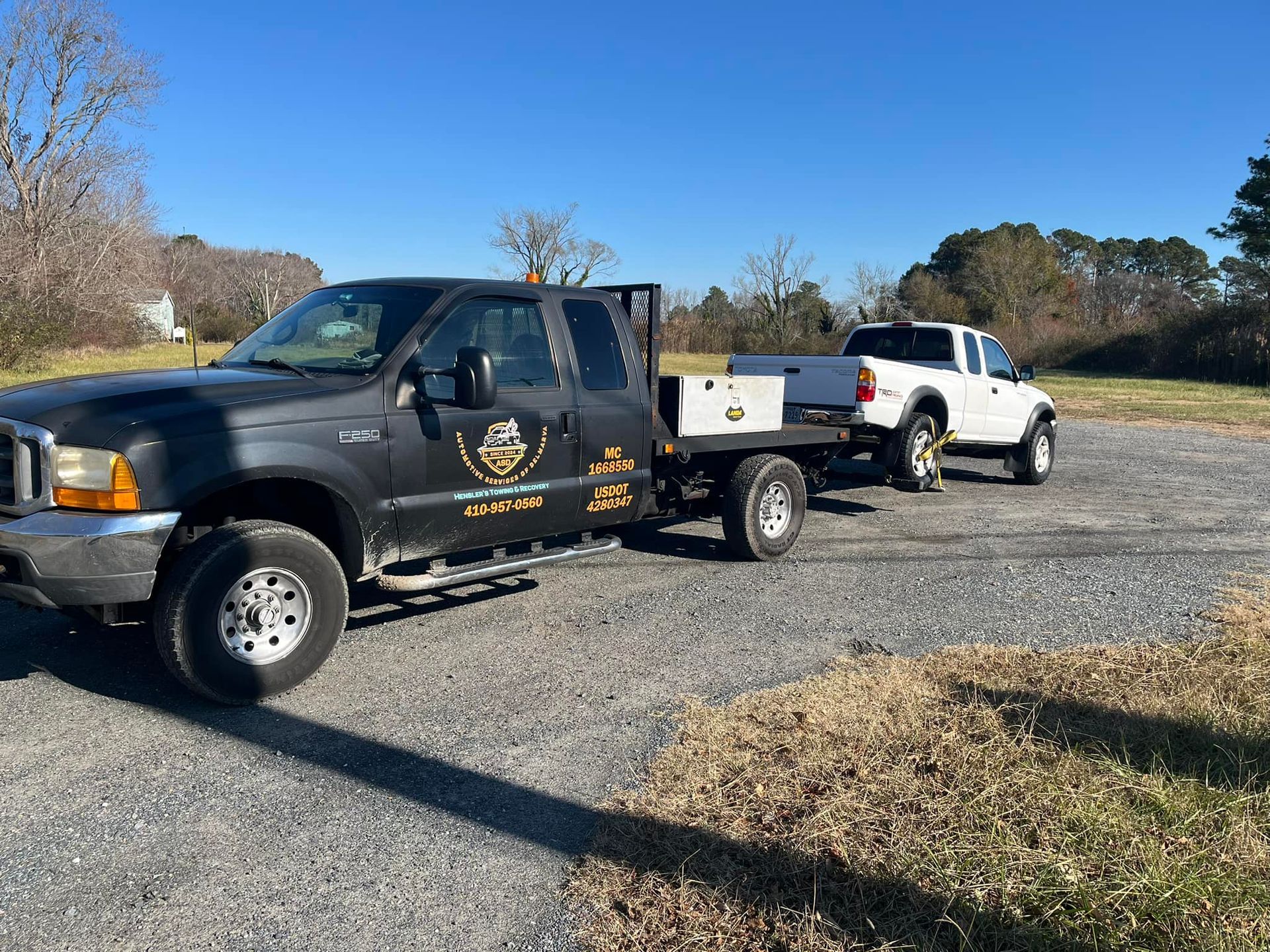 Black truck towing a white truck on a gravel driveway in a grassy area under a blue sky.