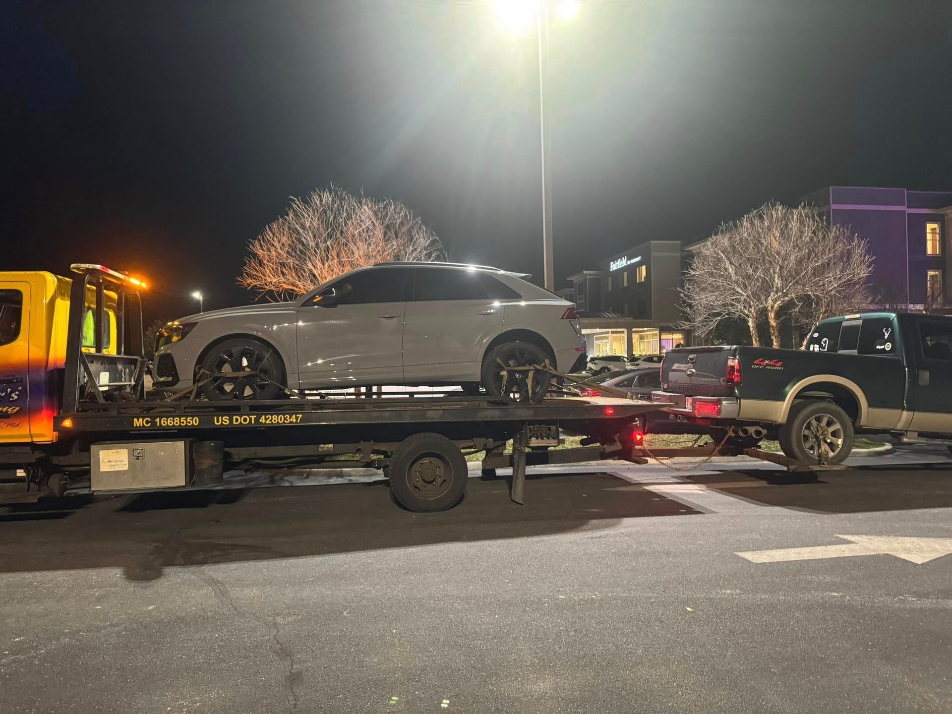 Gray car being towed on a flatbed truck at night, parked in a lot under a bright light.