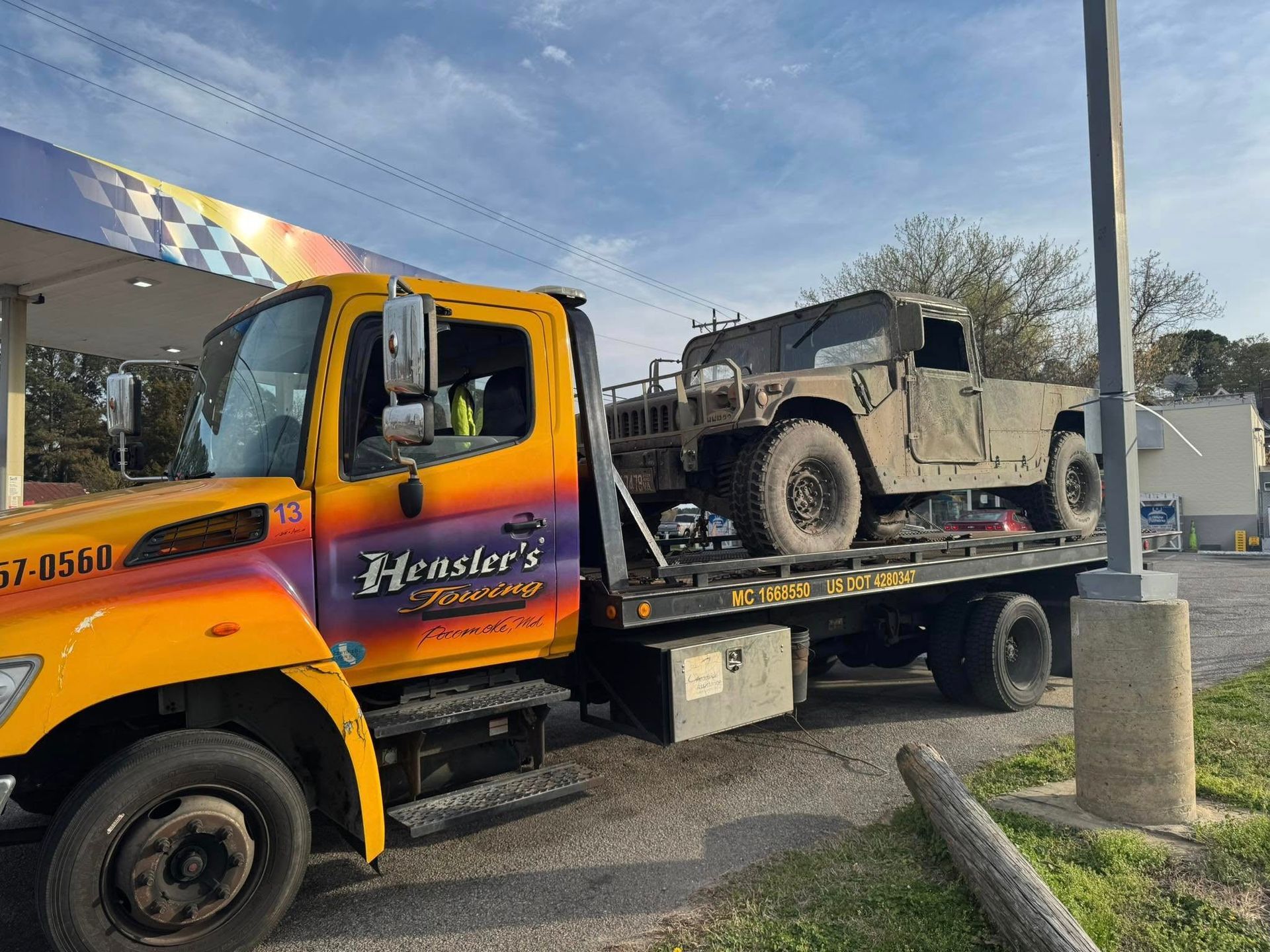 Yellow tow truck transporting a camouflage Humvee, parked near a gas station.