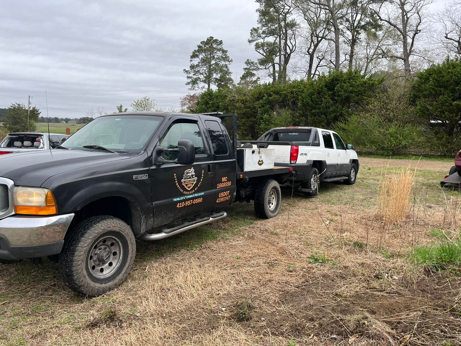 Black Ford pickup truck towing a white Chevrolet Avalanche flatbed truck in a grassy area.