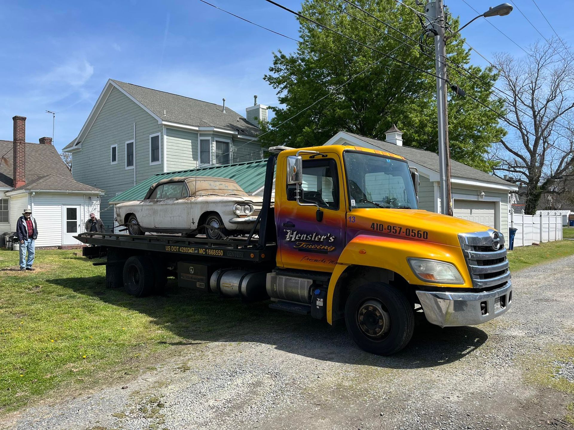 Tow truck with a white convertible parked on a residential street. Yellow and black truck, green house in the background.