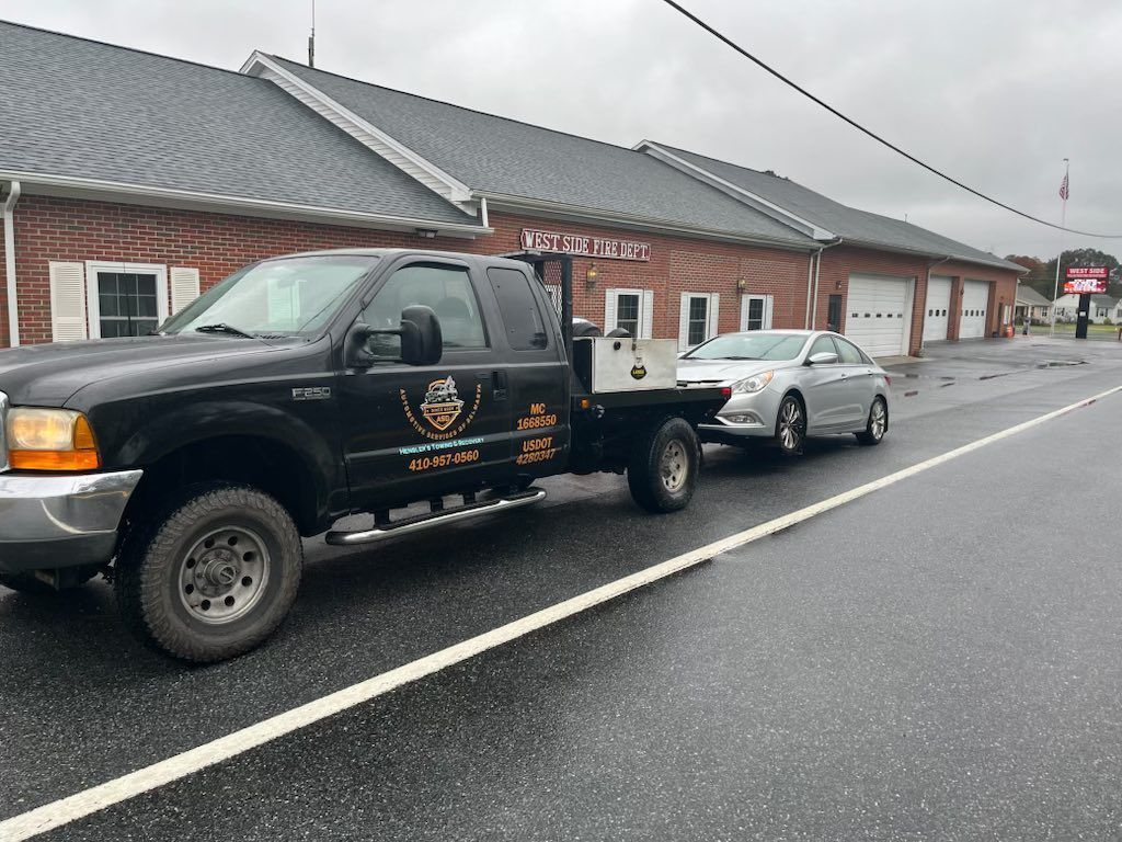 Black pickup truck towing a silver car on a wet road in front of a brick building.
