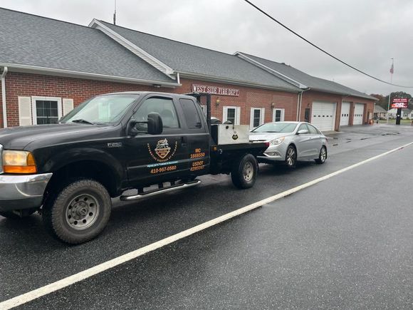 Black truck towing a silver car on a wet road in front of a brick building.