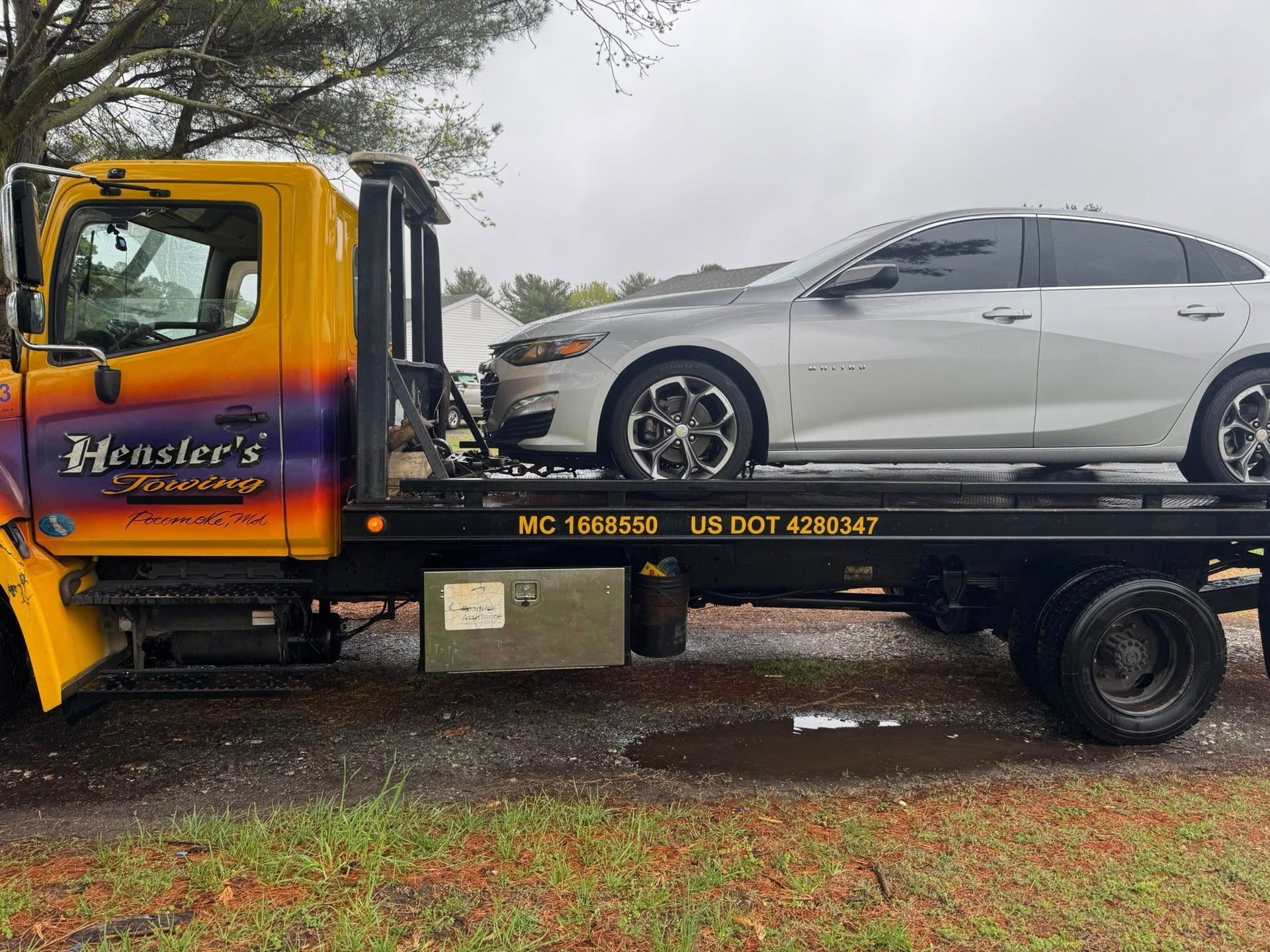 Silver car loaded onto a yellow tow truck. The truck is parked outdoors.