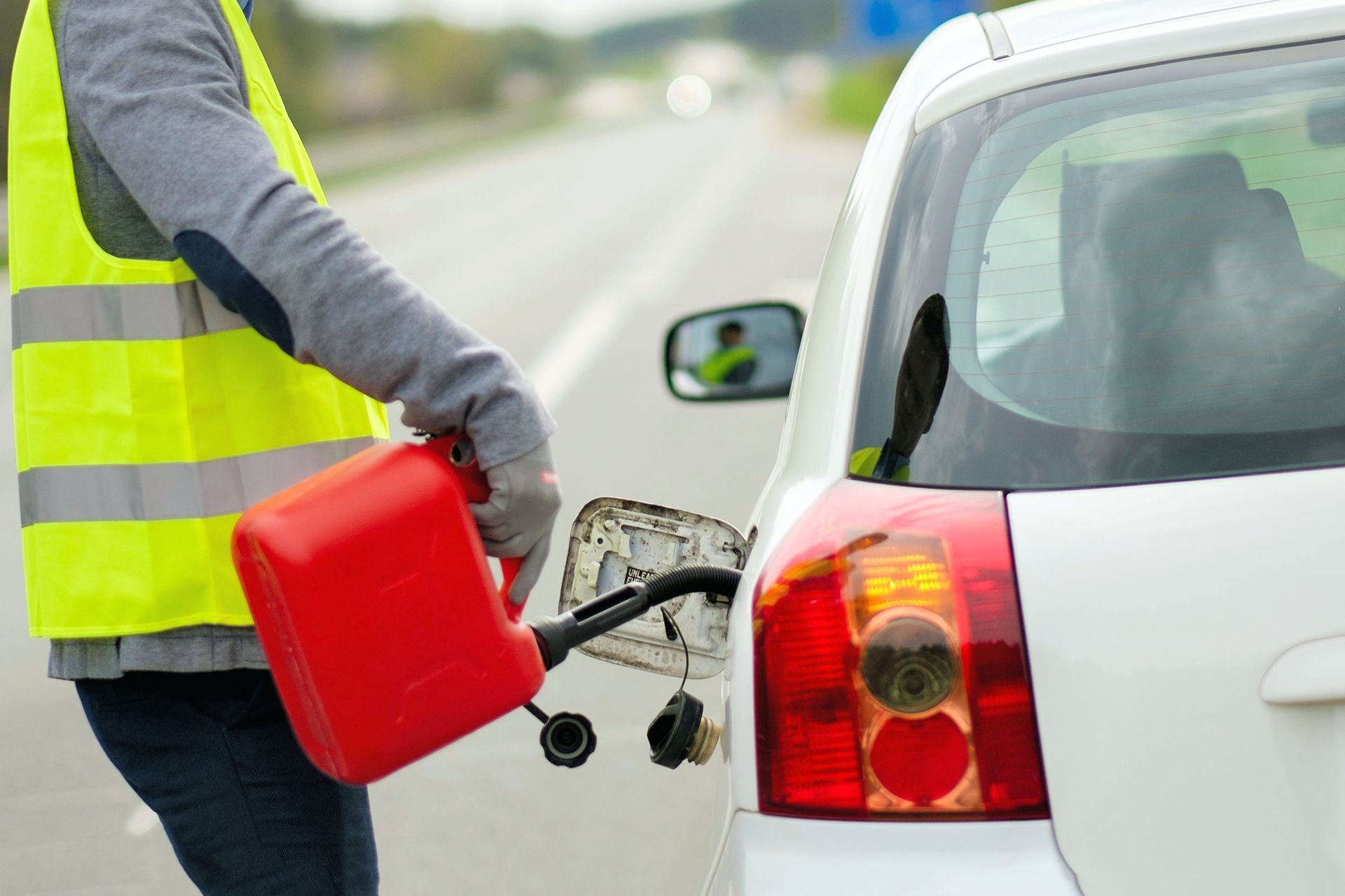 Person in safety vest refuels a white car with a red gas can on the side of a road.