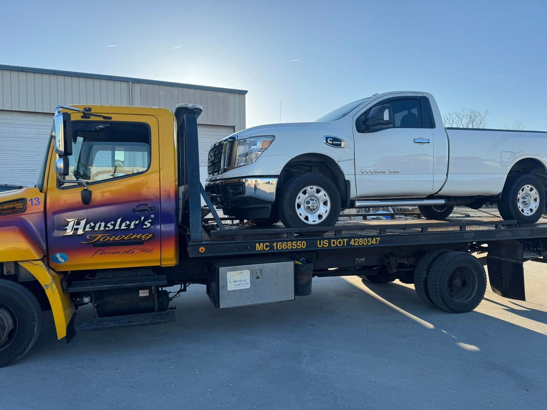 A white pickup truck being towed by a yellow Hensler's Towing truck on a sunny day.