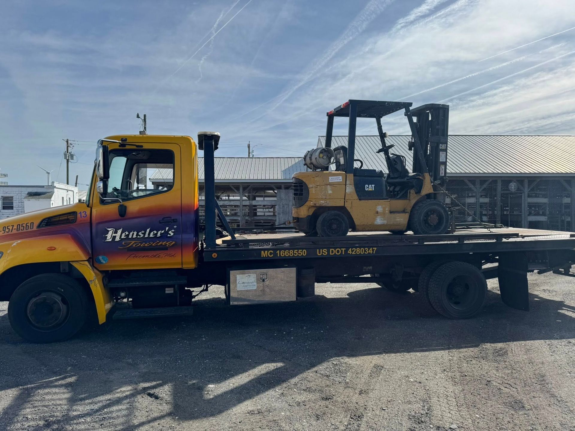 Yellow tow truck with a forklift loaded on its flatbed. Sunny outdoor setting.