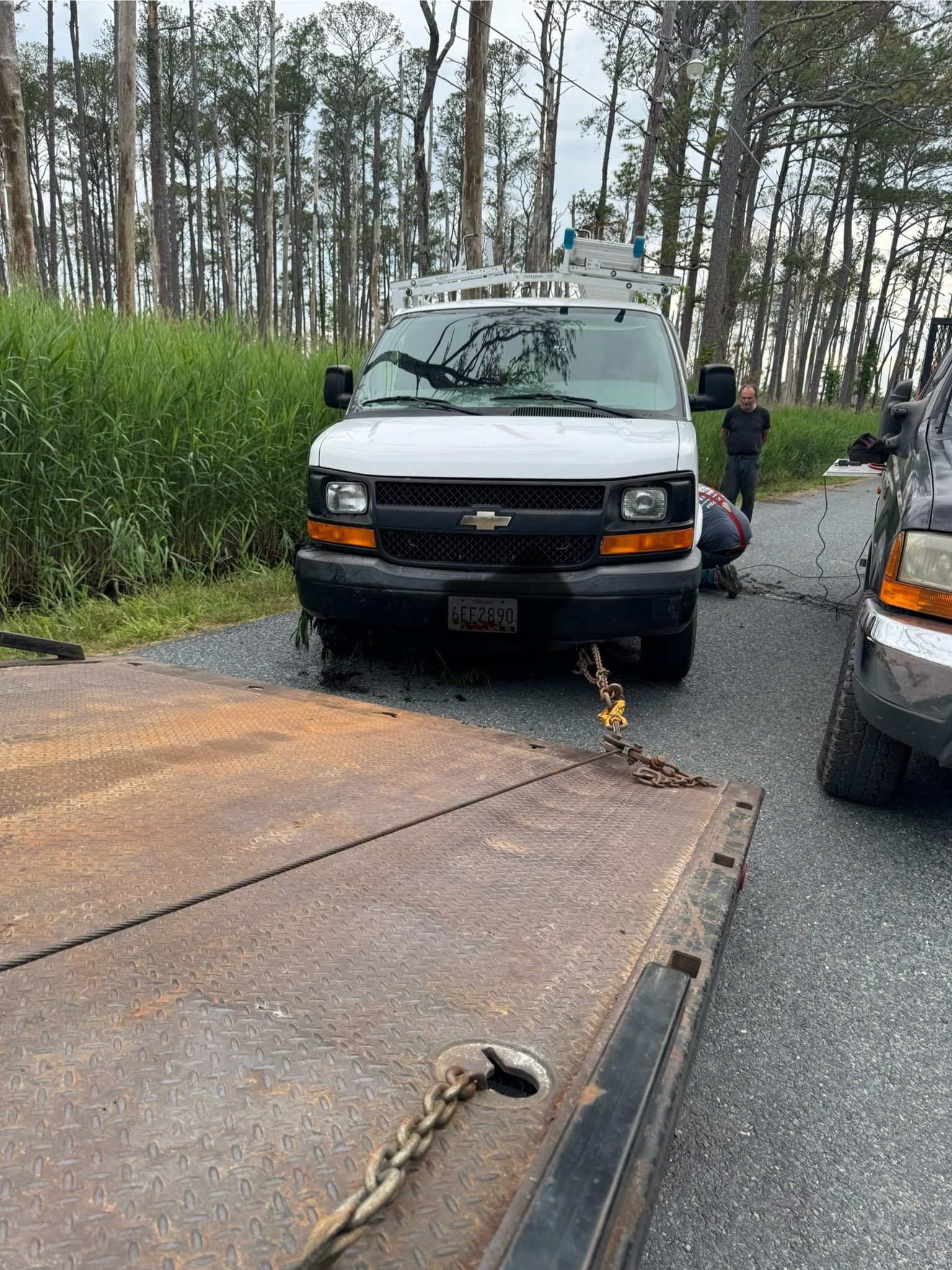 White van being loaded onto a rusty flatbed trailer, two people nearby.