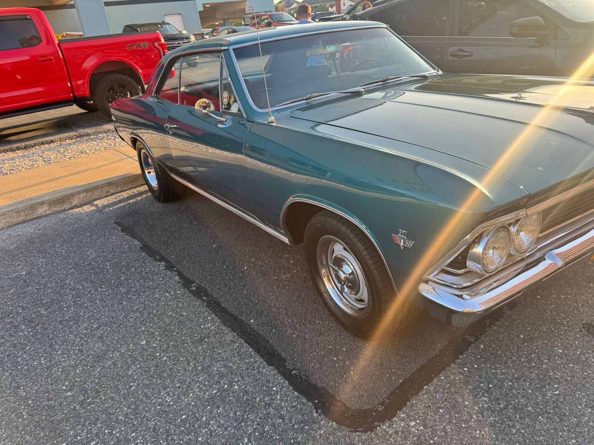Green classic car parked on asphalt with a red truck in the background.