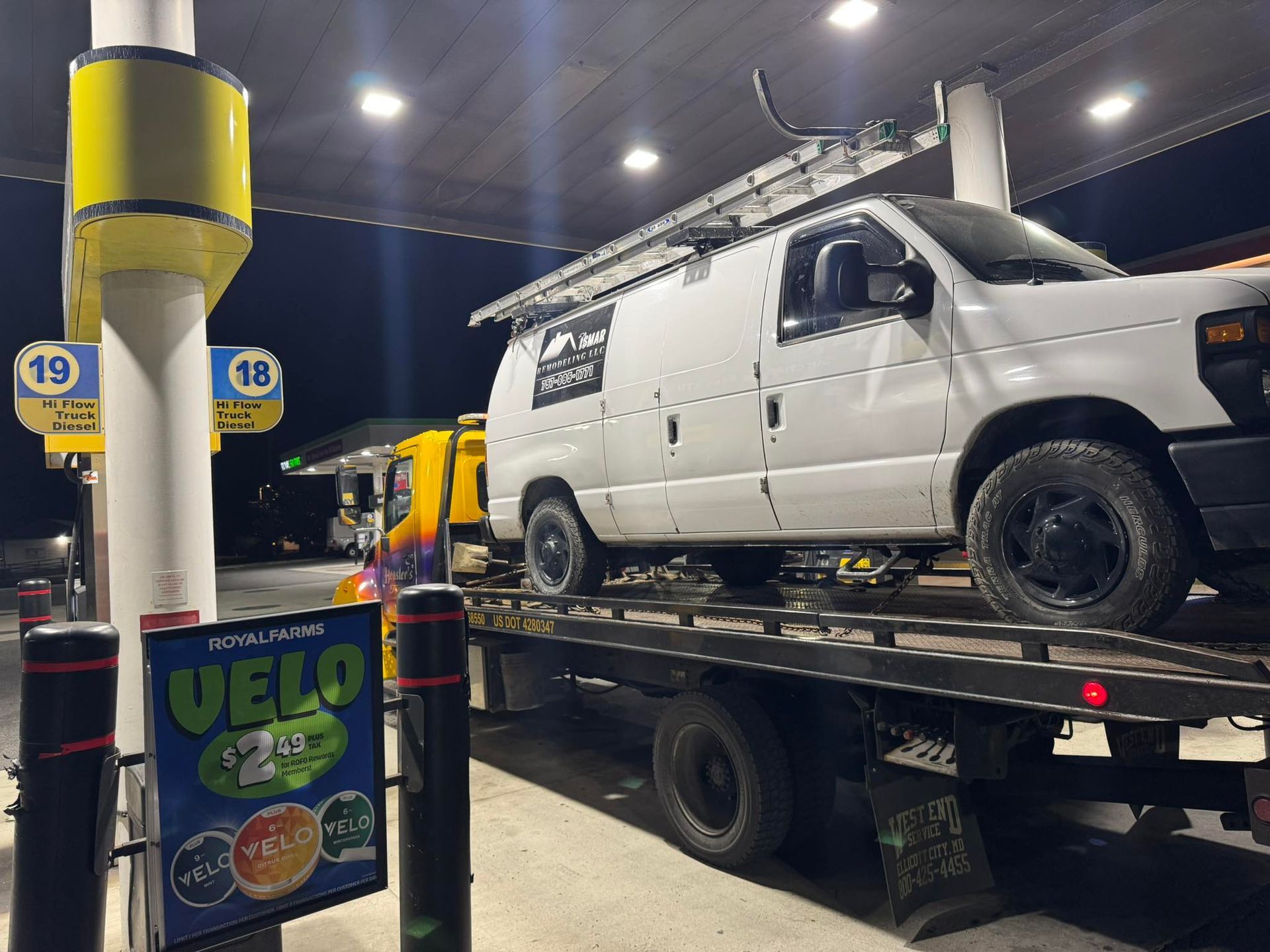 White van on a tow truck at a gas station at night. Sign shows fuel price. Yellow and black gas pumps.