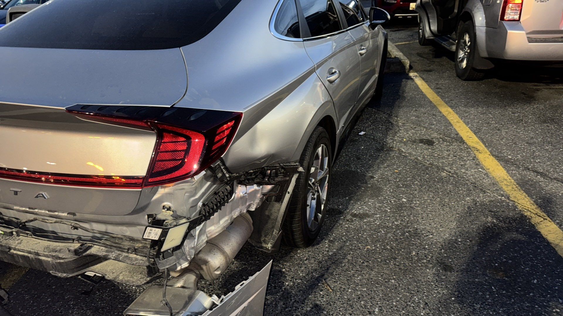 Damaged silver car rear end in parking lot; crumpled bumper, exposed undercarriage.