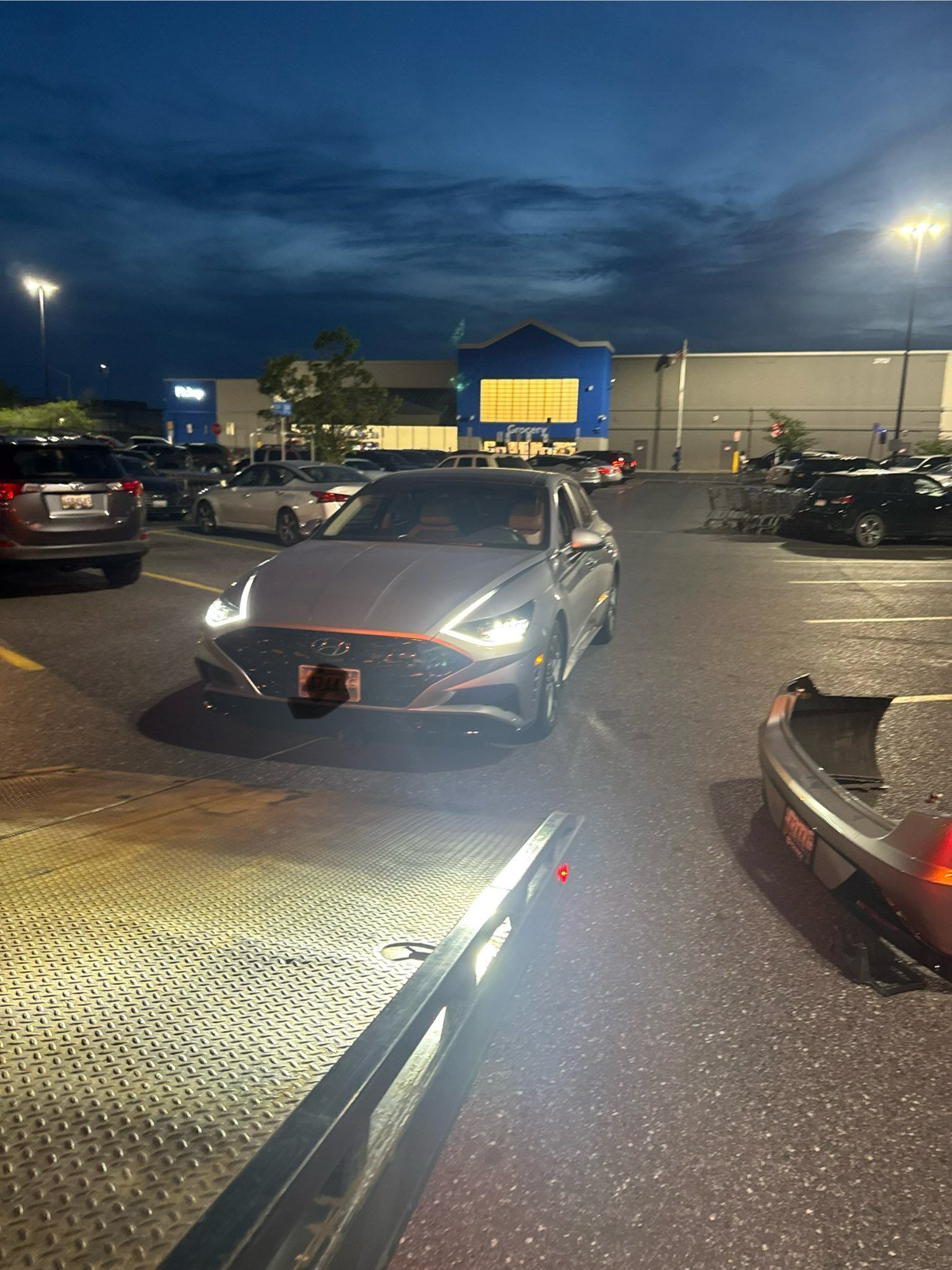 Silver sedan in a parking lot, headlights on, with a Walmart store in the background at dusk.