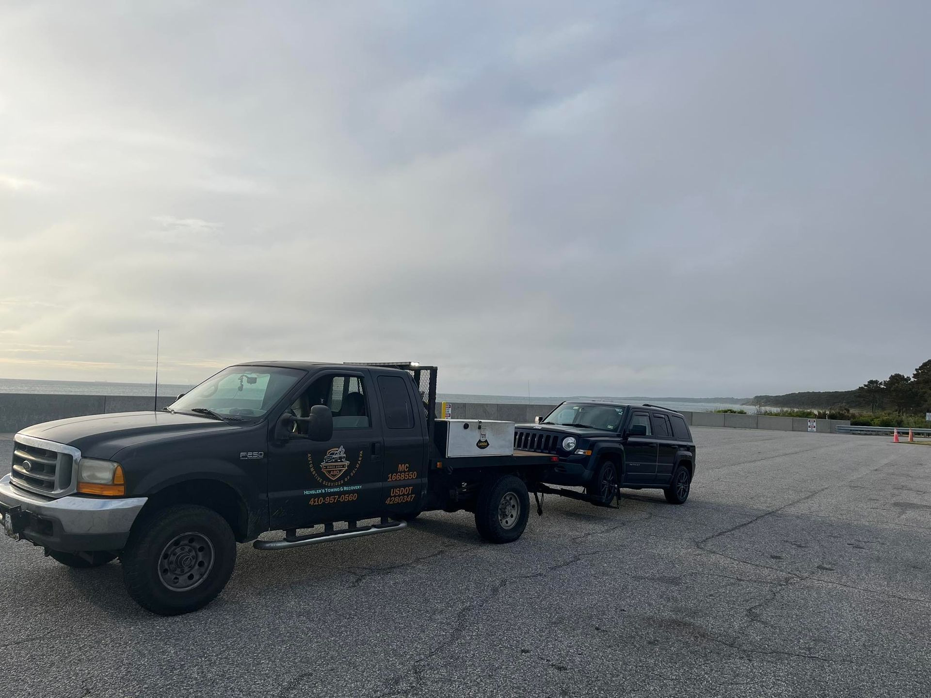 Black tow truck towing a black SUV on a gravel surface under a cloudy sky.