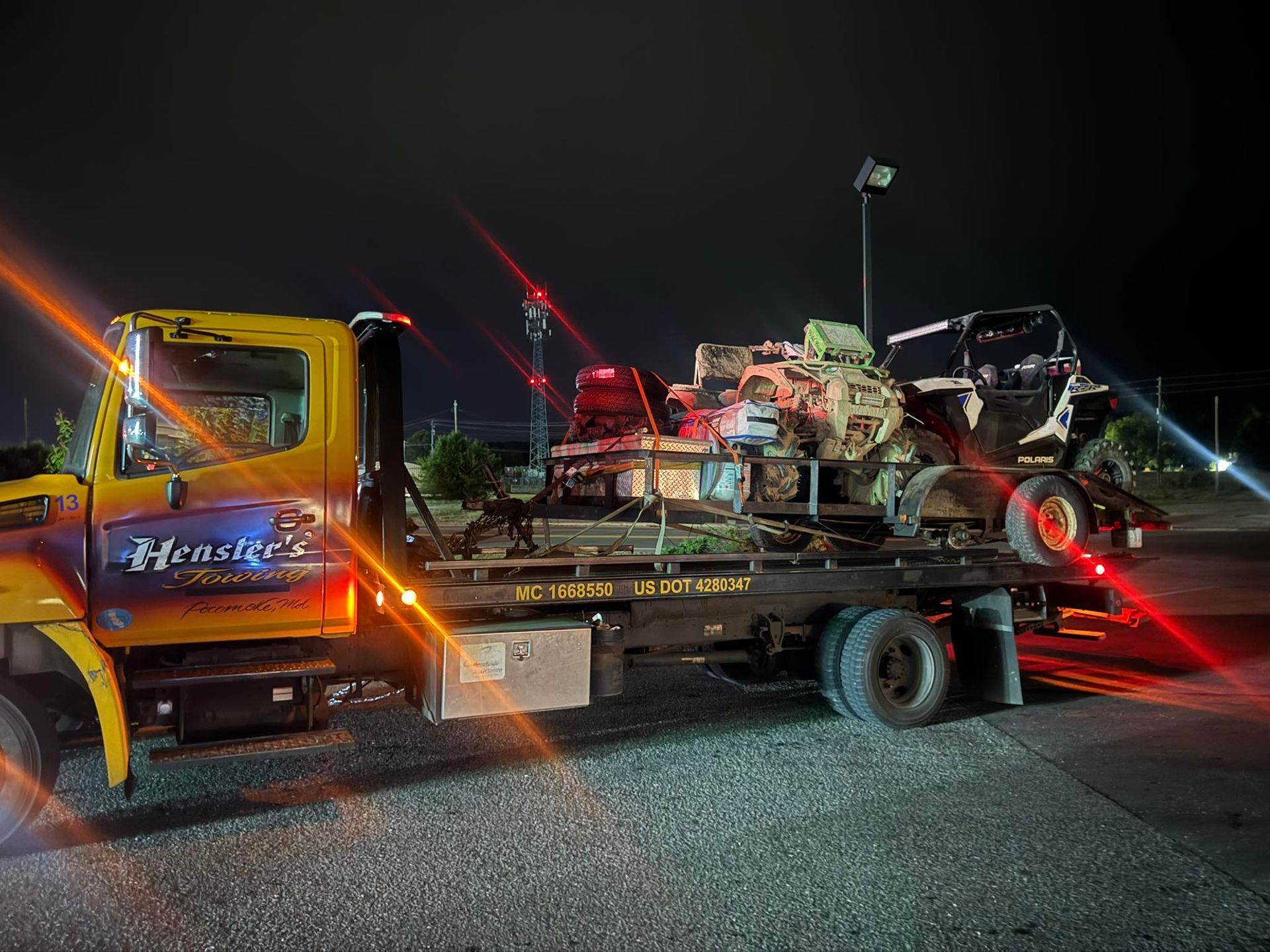 Yellow tow truck carrying a damaged vehicle at night; red and white lights visible.