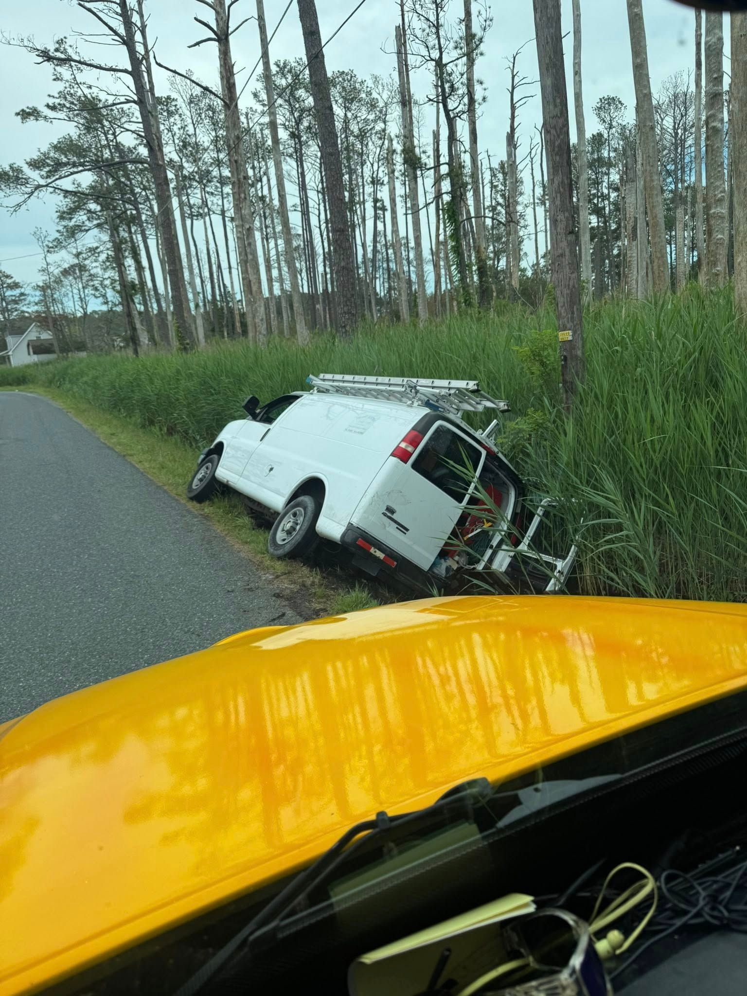 White work van off the road in tall grass next to a road, yellow vehicle in foreground.