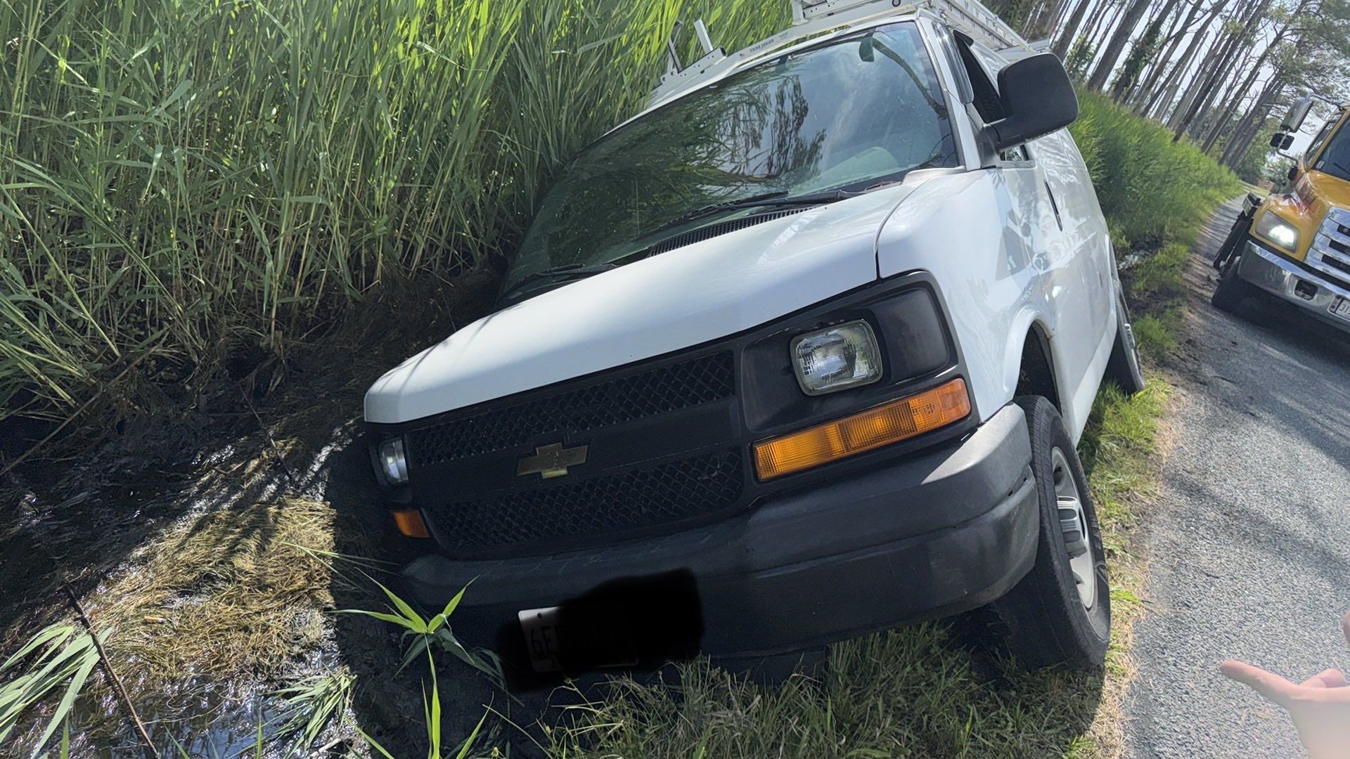 White van stuck in a ditch next to tall grass on the side of a road; a truck is visible behind it.
