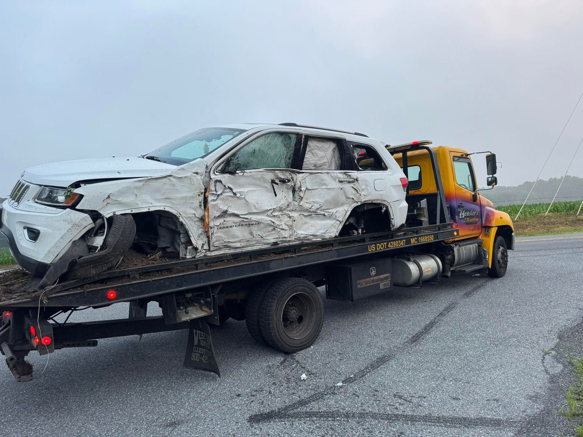 White SUV severely damaged, towed on a flatbed truck on a rural road.
