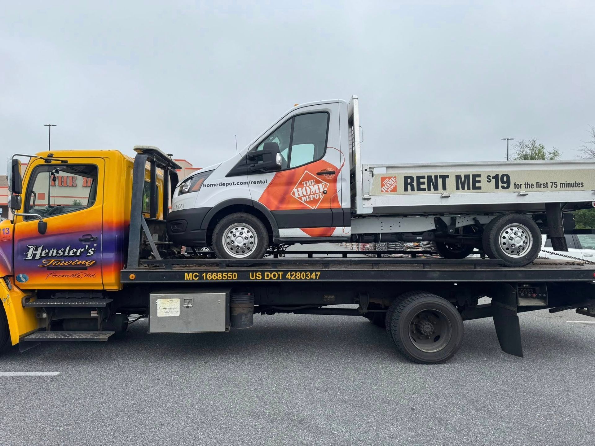 Yellow tow truck carrying a white and orange cargo van on a flatbed trailer.