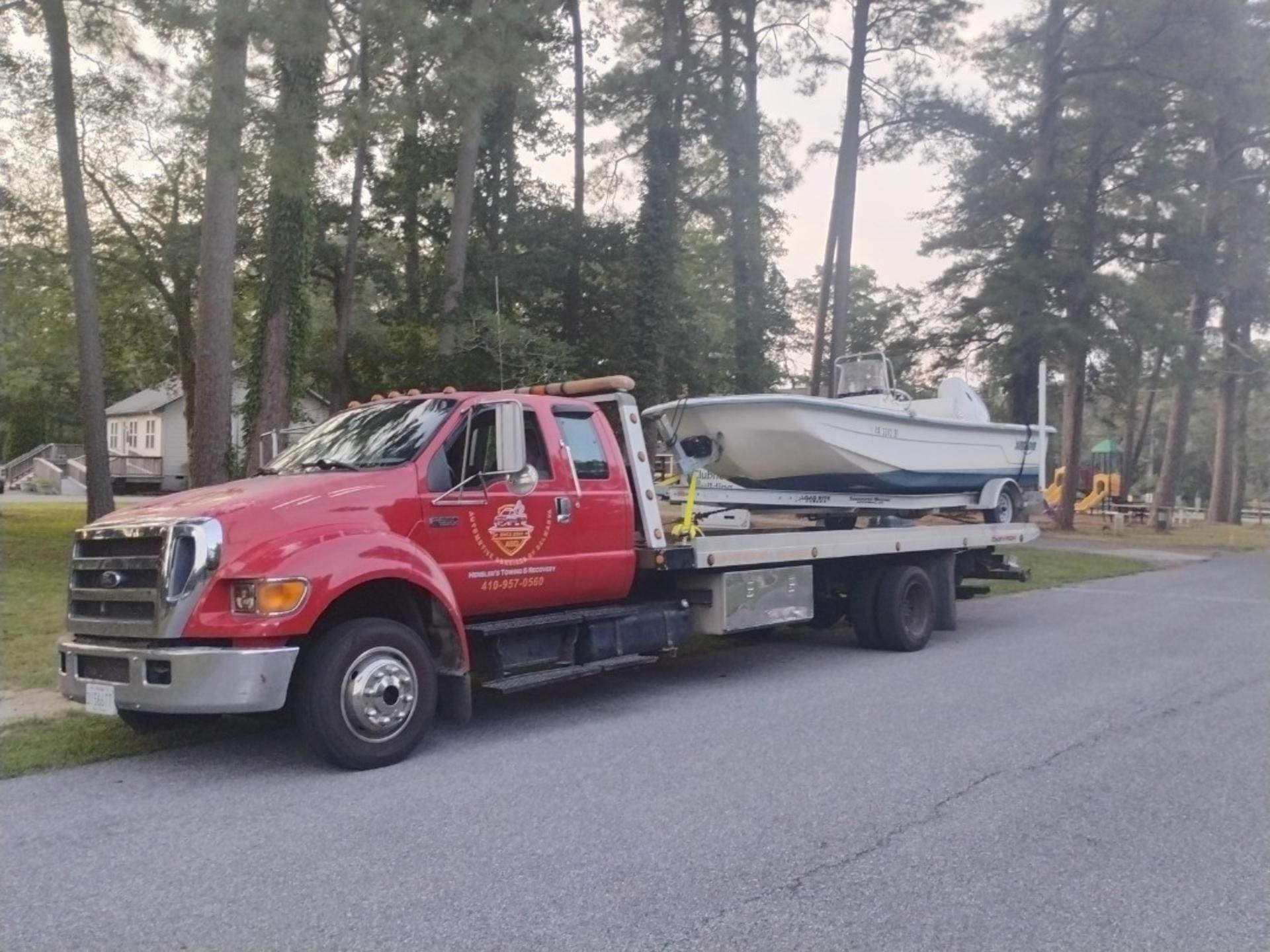 Red tow truck carrying a white boat on a flatbed, parked on a residential street.