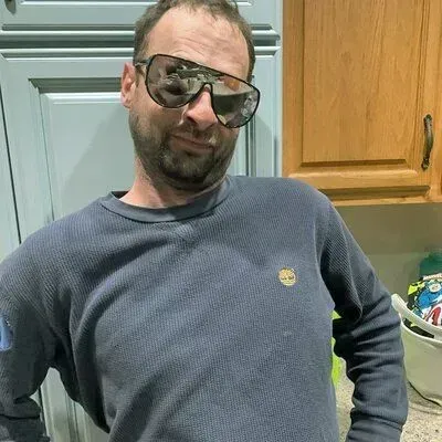 Man wearing sunglasses and a blue long-sleeved shirt, standing in front of kitchen cabinets.