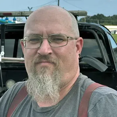 Man with glasses and goatee, wearing a gray shirt and suspenders, standing in front of a tow truck.
