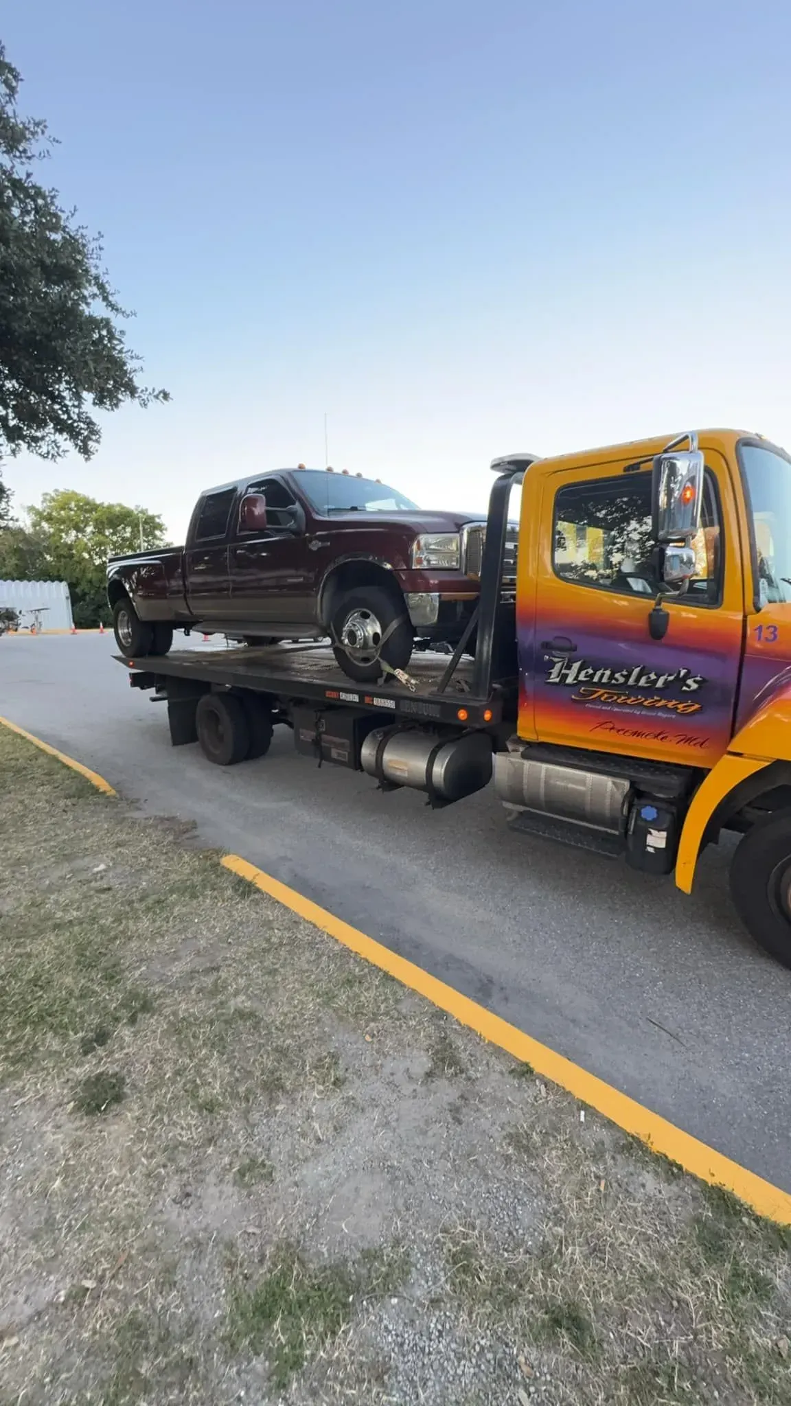 Tow truck transporting a maroon pickup truck on a paved road; the tow truck is yellow and has the business name on the side.