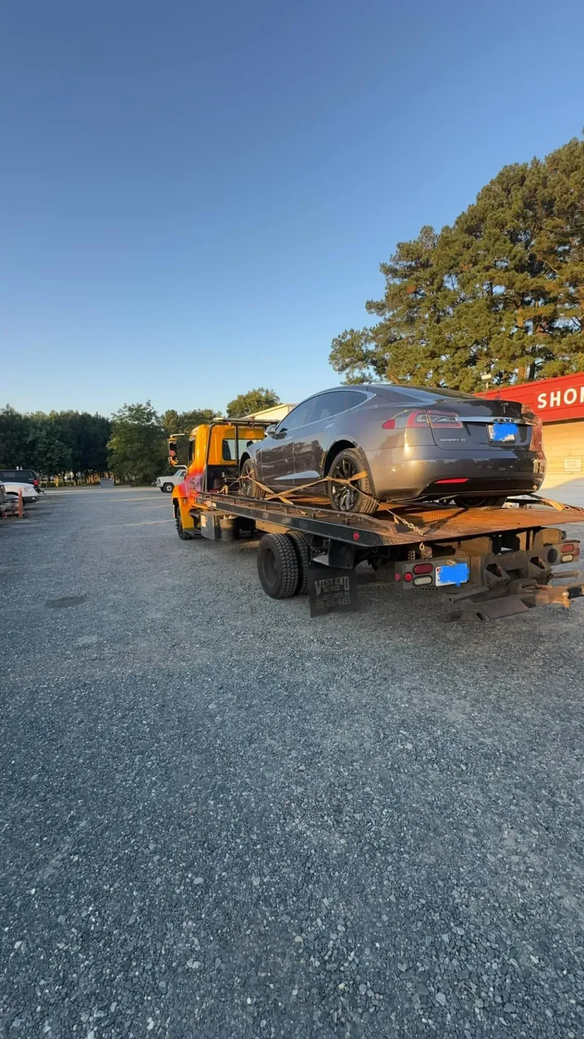 A gray car on a tow truck, parked on a gravel lot near a building under a blue sky.