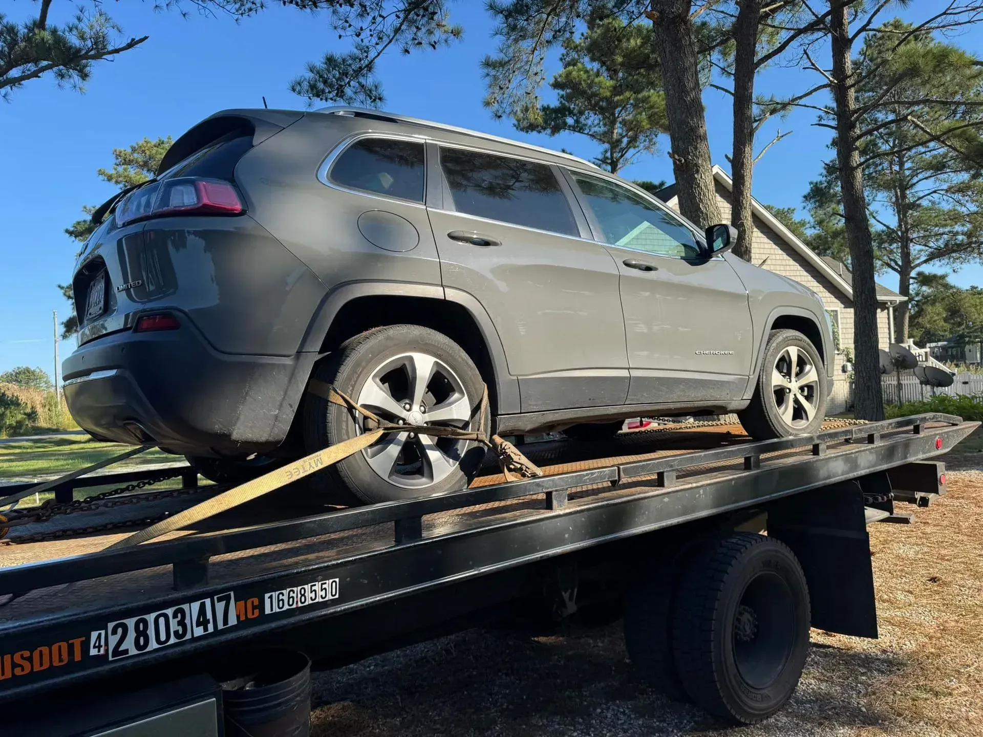 Gray Jeep Cherokee SUV being towed on a flatbed tow truck outdoors on a sunny day.