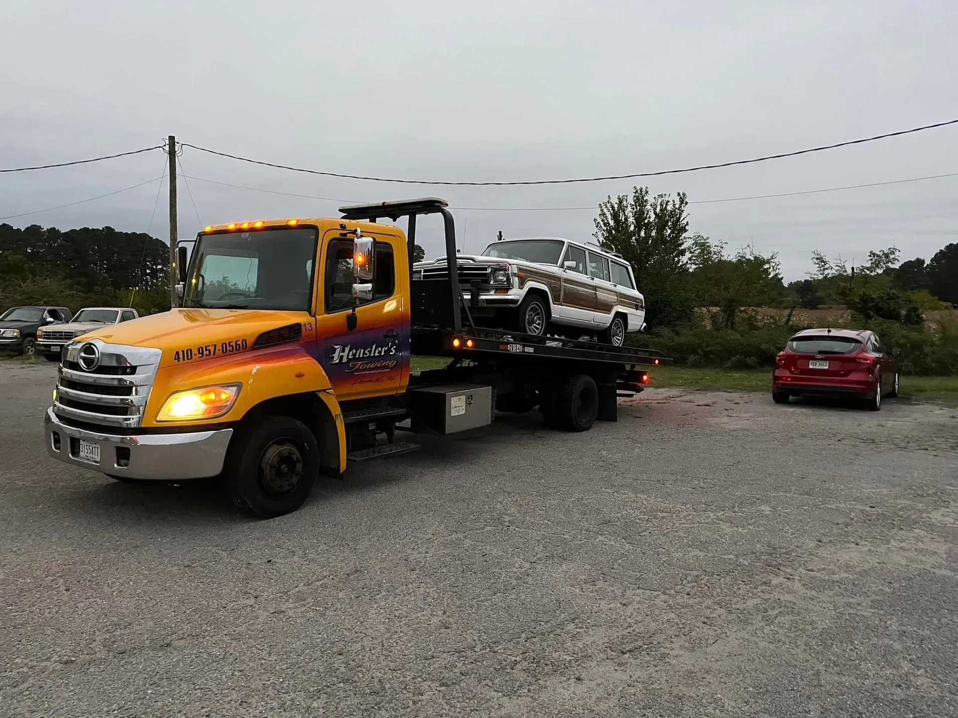 Yellow tow truck with a vintage Jeep on its flatbed, parked on gravel. A red car is to the right.
