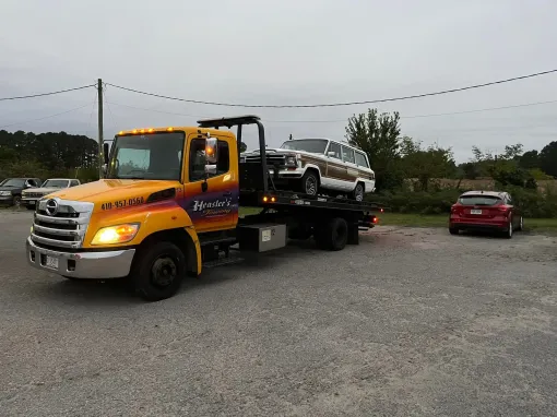 Yellow tow truck with a vintage Jeep on its flatbed, parked on gravel. A red car is to the right.