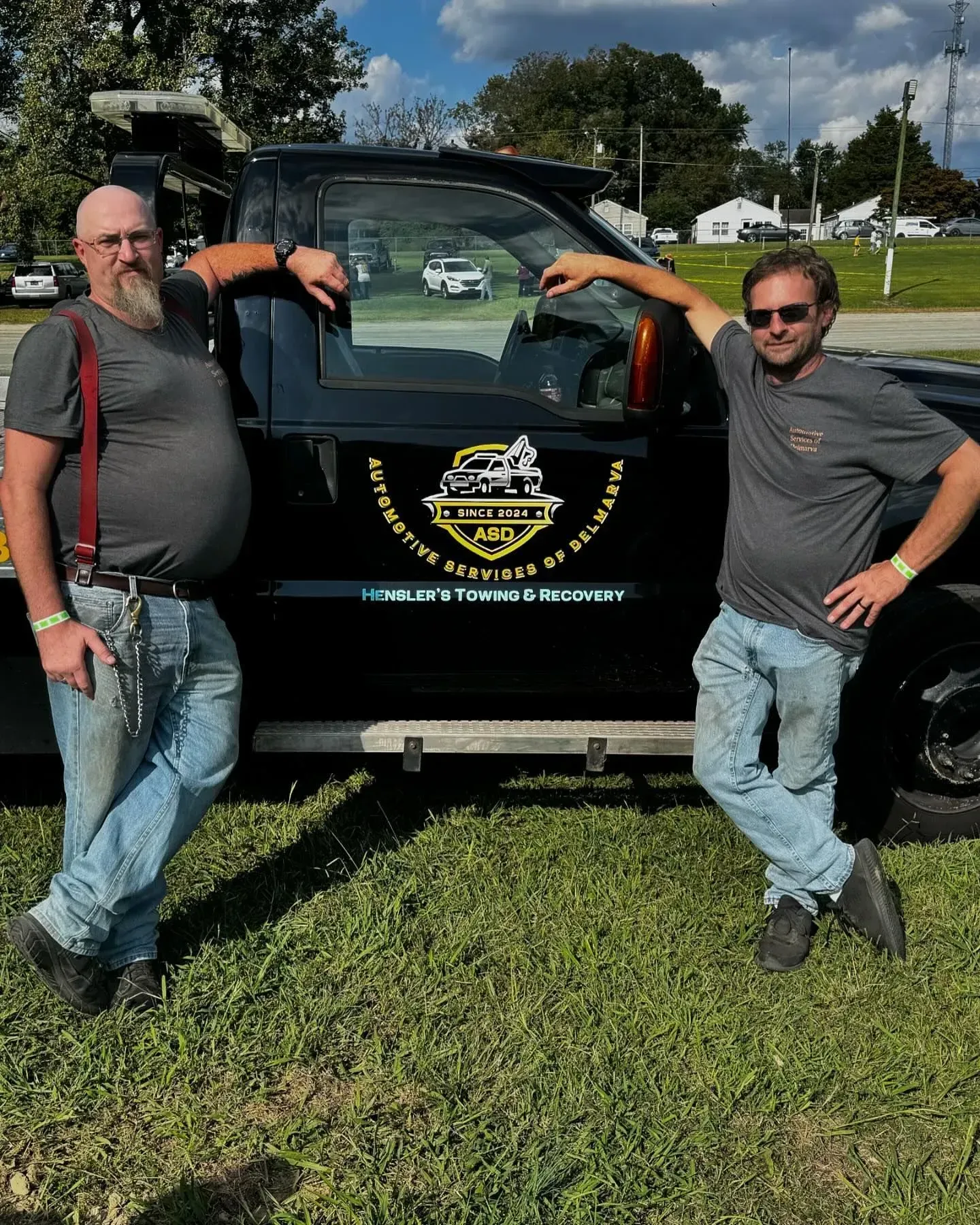 Two men lean on a black tow truck with logo. They're on grass outdoors. One has a beard, the other, sunglasses.