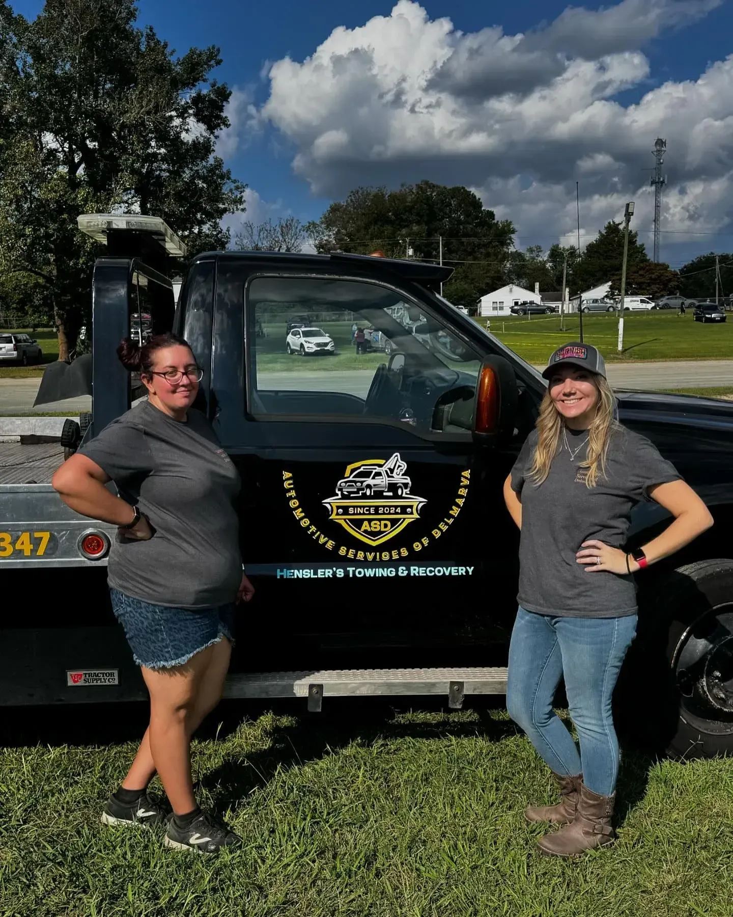 Two women standing next to a black tow truck with logo on side, outdoors under partly cloudy sky.