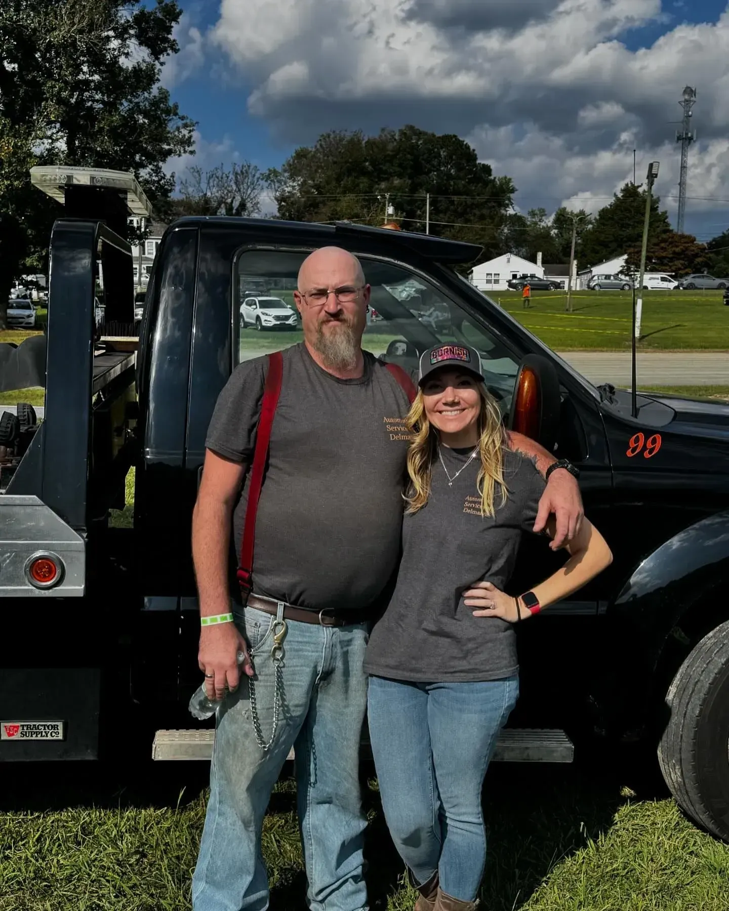 A man and woman stand by a black tow truck in a field, smiling.