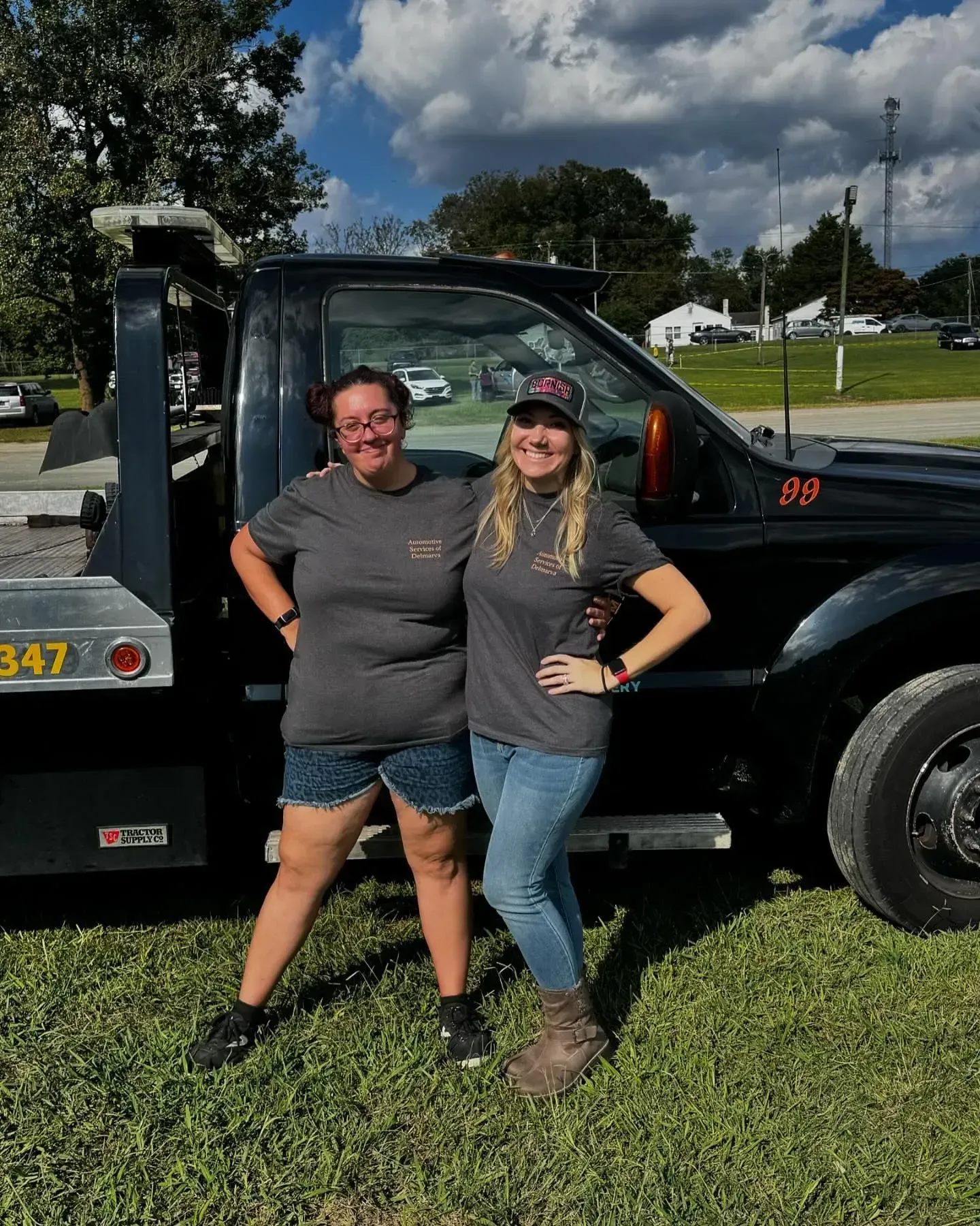 Two women stand smiling next to a black tow truck on a grassy area, wearing matching t-shirts.