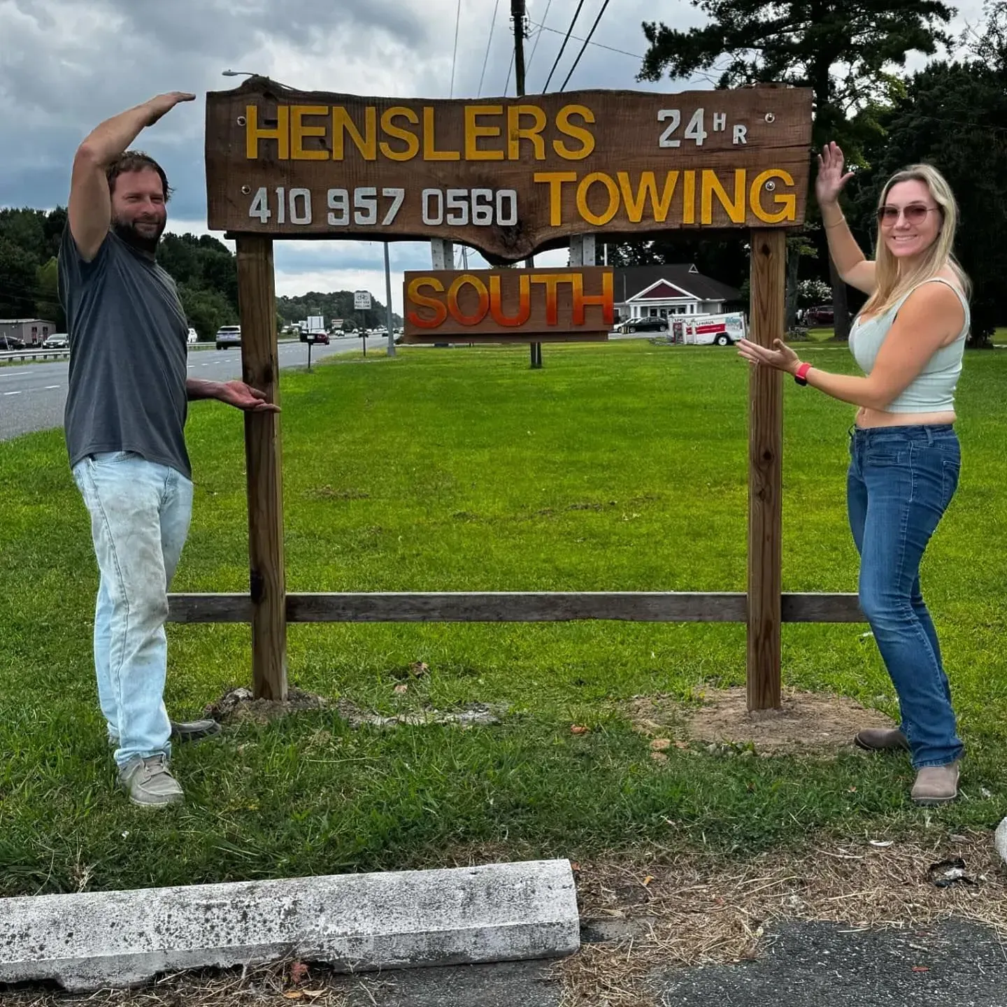 Two people stand next to a Hensler's Towing sign. The man is on the left, woman on right, and both are holding up their arms.