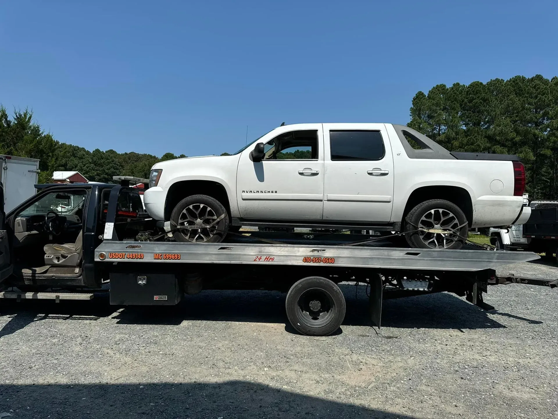 White Chevrolet Avalanche truck on a flatbed tow truck in a junkyard on a sunny day.