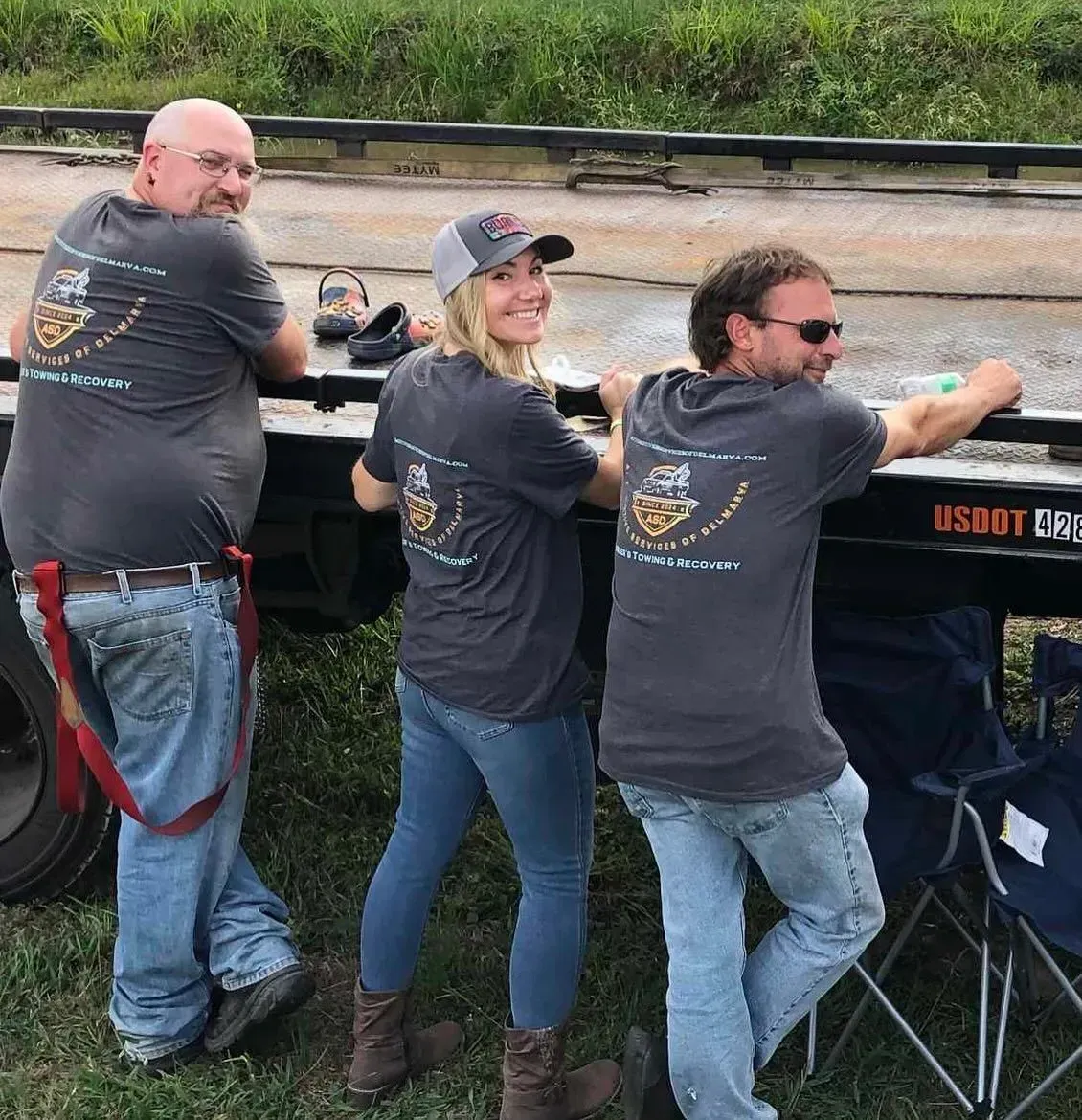 Three people wearing matching t-shirts lean on a trailer. They smile outdoors.