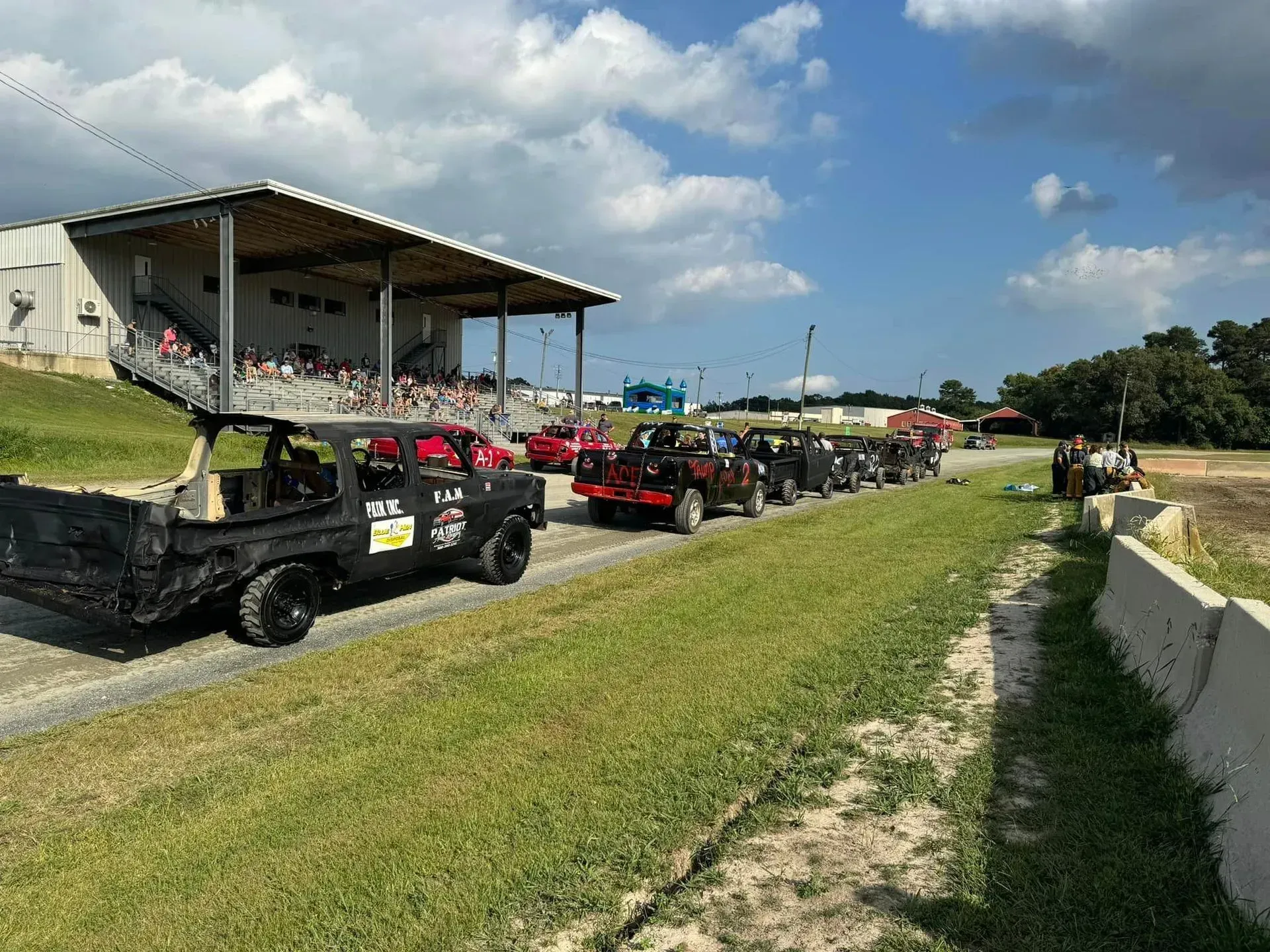Vehicles lined up on a dirt track, with a viewing stand and spectators. Sunny day.