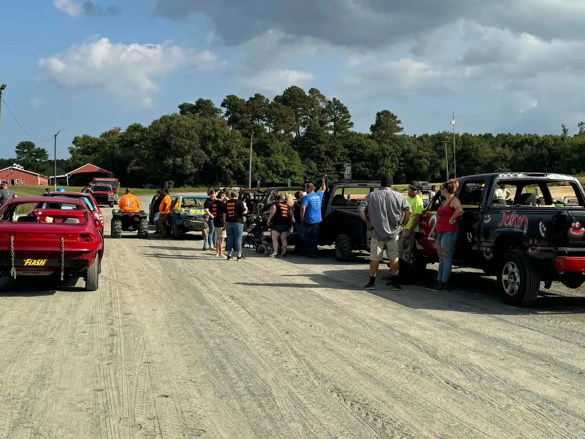 Cars and people gather on a dirt road, likely at a race or event; sunny day.