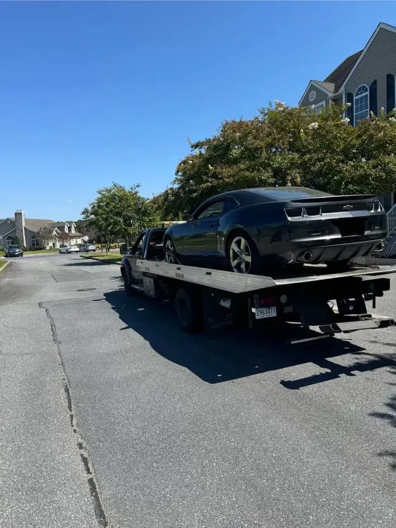 Black Camaro on a flatbed tow truck on a residential street under a blue sky.
