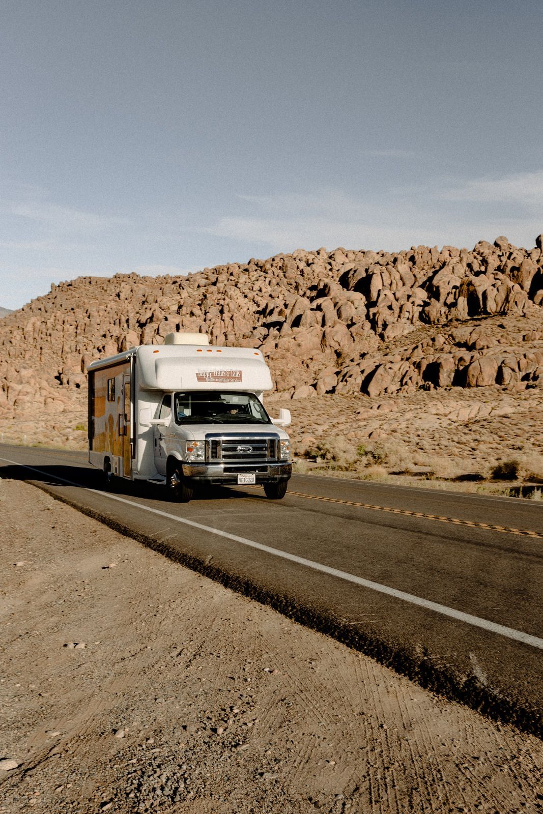 RV driving down a road in a desert landscape with rocky hills under a clear blue sky.