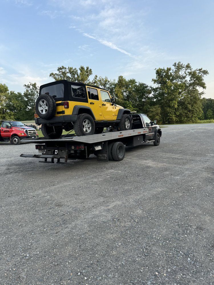 Yellow Jeep being transported on a flatbed tow truck outdoors on a gravel lot.