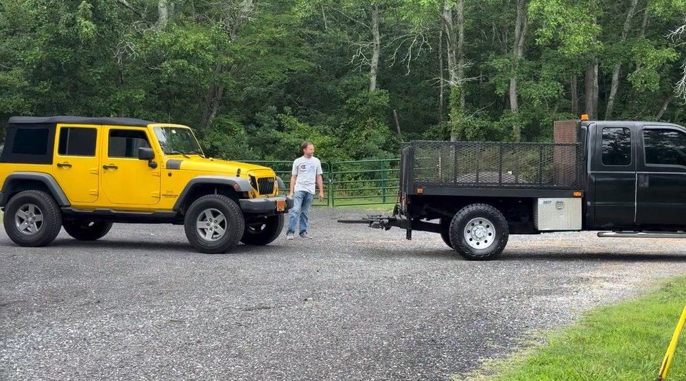 Yellow Jeep and black pickup truck pulling an empty trailer on a gravel road, person standing nearby.