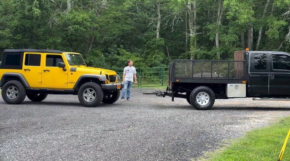 Yellow Jeep and black truck towing a trailer on a gravel driveway, person standing nearby.
