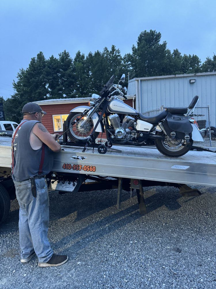 Man securing motorcycle on tow truck bed. Gray motorcycle with black saddlebags. Outdoor setting.